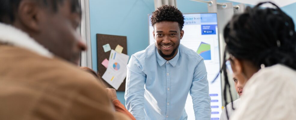 A portrait of a sales representative leaning against a table, framed by two coworkers discussing the presentation he just gave